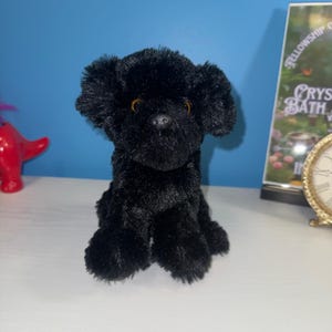 Puede incluir: Un peluche con forma de perro con pelaje negro, orejas caídas y ojos de color ámbar. El juguete está en posición sentada con una pequeña nariz negra y una apariencia suave y texturizada, diseñado como una figura de peluche coleccionable.