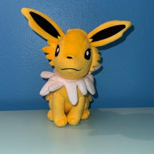 Puede incluir: Un peluche amarillo de una criatura con grandes orejas puntiagudas con puntas negras. Tiene un cuello blanco con volantes y ojos negros. El juguete está sobre una superficie azul claro.