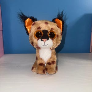 May include: A plush toy lynx with large, glittery orange eyes, a black nose, and a white chest. The lynx has brown spots and tufted ears with black and orange accents. It is sitting upright on a white surface against a blue background.
