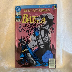 Puede incluir: Portada de cómic de Detective Comics con Batman. La portada tiene un fondo rosa con una ilustración de Batman y Bane. El texto "BATMAN" está en letras amarillas grandes.
