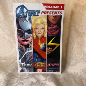 Op de afbeelding: A-Force Presents Volume 1 stripboek. De cover bevat illustraties van Thor, Captain Marvel en Ms. Marvel. Het stripboek heeft een witte achtergrond met de titel in blauw en rood. Het Marvel Comics-logo is zichtbaar.