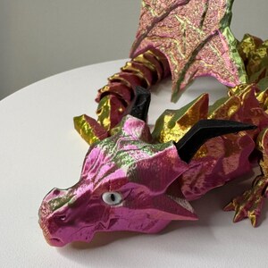 May include: A pink and gold dragon figurine with iridescent scales and black horns. The dragon is posed with its head turned to the side and its wings spread out.