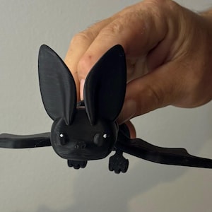 May include: A black 3D printed bat figurine with large ears and white eyes. The bat is posed in flight with its wings spread out.