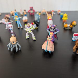 Puede incluir: Una colecci&oacute;n de peque&ntilde;as y coloridas figuras de personajes de Toy Story. Las figuras incluyen a Buzz Lightyear, Woody, Bo Peep y otros. Los juguetes est&aacute;n dispuestos sobre una superficie oscura.