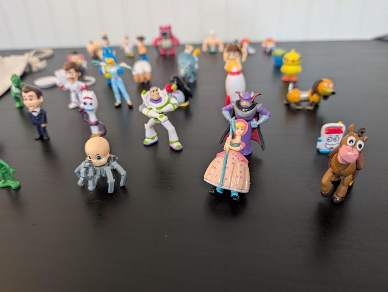 Puede incluir: Una colecci&oacute;n de peque&ntilde;as y coloridas figuras de personajes de Toy Story. Las figuras incluyen a Buzz Lightyear, Woody, Bo Peep y otros. Los juguetes est&aacute;n dispuestos sobre una superficie oscura.
