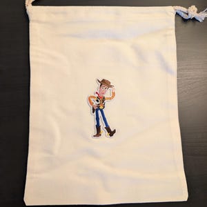 Puede incluir: Una bolsa con cord&oacute;n de color crema con una imagen de dibujos animados colorida de Woody de Toy Story. La bolsa tiene un dise&ntilde;o simple con un cierre de cord&oacute;n de cuerda. La imagen de Woody est&aacute; en el centro de la bolsa.