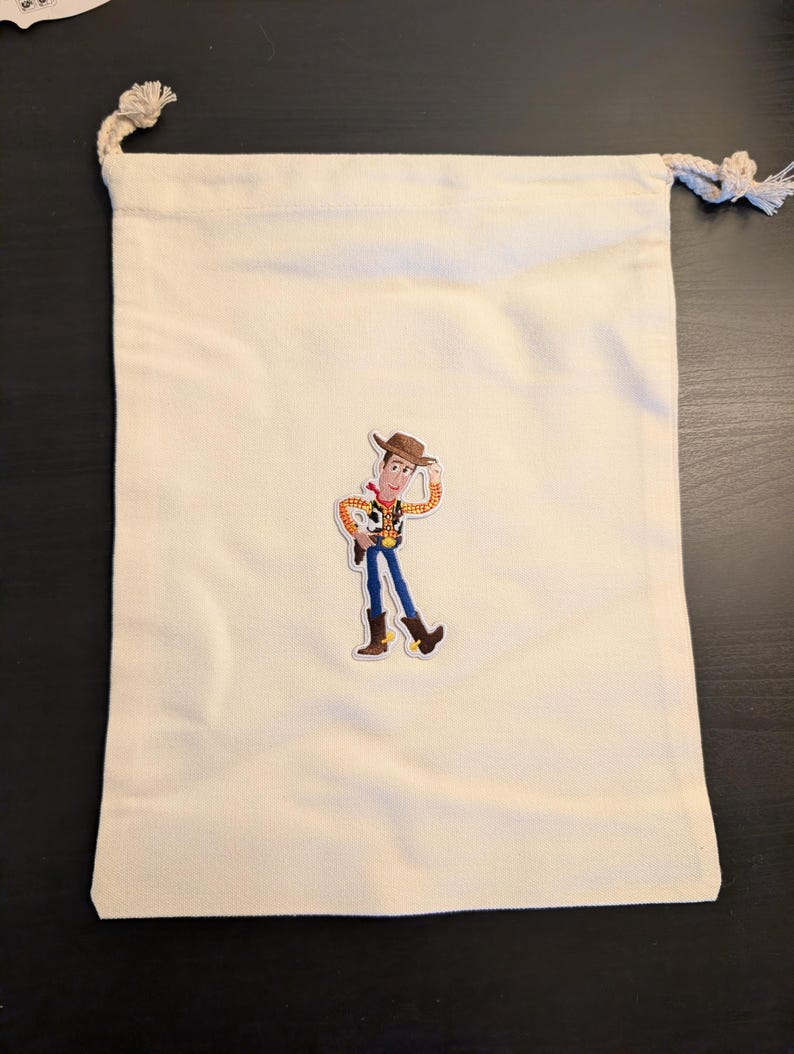 Puede incluir: Una bolsa con cord&oacute;n de color crema con una imagen de dibujos animados colorida de Woody de Toy Story. La bolsa tiene un dise&ntilde;o simple con un cierre de cord&oacute;n de cuerda. La imagen de Woody est&aacute; en el centro de la bolsa.