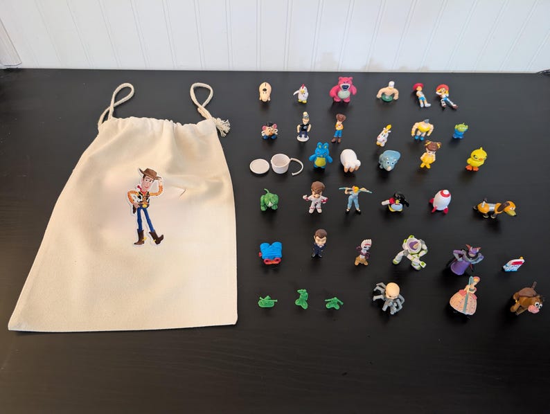 Puede incluir: Una colecci&oacute;n de figuras de personajes de Toy Story y una bolsa con cord&oacute;n. La bolsa es de color crema con un gr&aacute;fico del personaje Woody. Las figuras son de varios tama&ntilde;os y colores, con personajes como Buzz Lightyear y Rex.