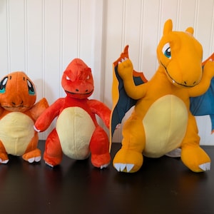 May include: Three plush toys featuring a small orange bipedal creature with a cream belly, a medium red bipedal creature with a cream belly, and a large orange winged creature with a cream belly, blue wing interiors, and white claws.