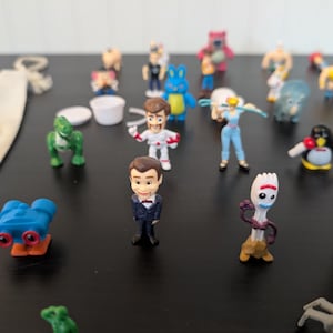 Puede incluir: Una colecci&oacute;n de figuras de personajes en miniatura, que incluyen un dinosaurio verde, un ping&uuml;ino y una figura con un traje espacial. Los juguetes est&aacute;n colocados sobre una superficie oscura, con algunos otros objetos en el fondo.