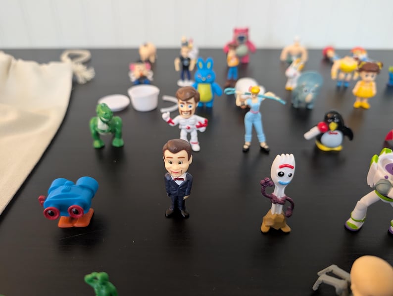 Puede incluir: Una colecci&oacute;n de figuras de personajes en miniatura, que incluyen un dinosaurio verde, un ping&uuml;ino y una figura con un traje espacial. Los juguetes est&aacute;n colocados sobre una superficie oscura, con algunos otros objetos en el fondo.