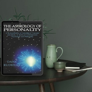 Könnte beinhalten: Ein Tablet zeigt das Buchcover von "The Astrology of Personality" von Dane Rudhyar. Das Cover zeigt ein blau-weißes Himmelsdesign. Neben dem Tablet stehen ein grüner Keramikkrug, eine kleine Tasse und ein Notizbuch auf einem dunklen Holztisch.