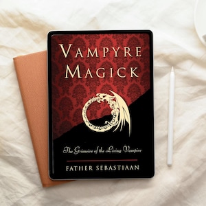 May include: A digital tablet displaying the book cover "Vampyre Magick" with a red damask pattern background and a gold dragon illustration. The book is next to a brown notebook and a white stylus.