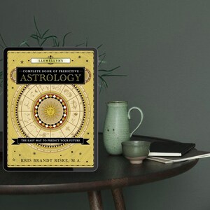 May include: A digital tablet displaying the cover of "Llewellyn's Complete Book of Predictive Astrology." The cover is gold and black with astrological symbols and the text "The Easy Way to Predict Your Future." A green ceramic pitcher and cup are on a dark wooden table.