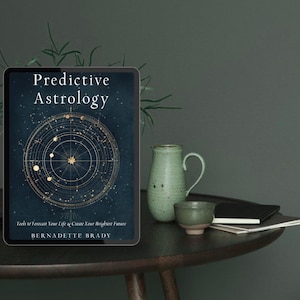 May include: A tablet displaying the book "Predictive Astrology" with a dark blue cover and a gold astrological chart design. Beside it are a green ceramic pitcher and a small bowl, with a black notebook on a dark wooden table.