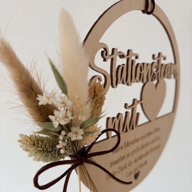 May include: A wooden circular sign with the words "Stationstraum mit" cut out. The sign features a heart cutout and is adorned with dried flowers and a brown ribbon. The sign has a natural wood color.