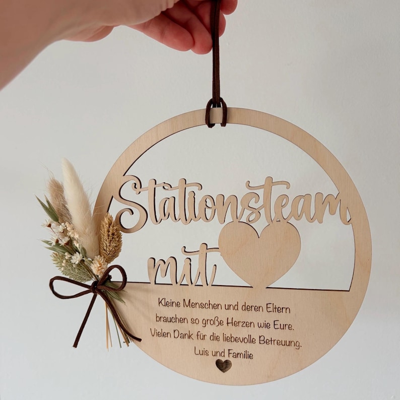 May include: A round wooden sign with the words "Stationsteam mit" cut out. The sign has a heart shape and a small bouquet of dried flowers and a brown ribbon. The sign also has text in German.