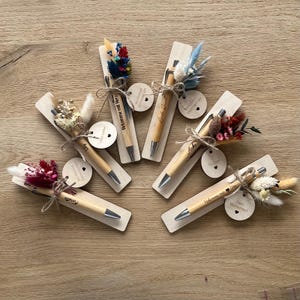 Könnte beinhalten: Sechs Holzstift-Halter mit getrockneten Blumen und einem Stift. Jeder Halter hat ein kleines Holzschild mit einer personalisierten Nachricht. Die Nachrichten lauten "Welcome Home", "Dein Wunschschild", "Mit Liebe", "Fin", "Arka" und "Welcome mit Liebe".