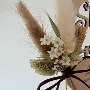 May include: A wooden wedding cake topper featuring a bouquet of dried flowers in beige, cream, and green hues. The bouquet is tied with a brown ribbon. The wooden base has decorative cutouts and text.