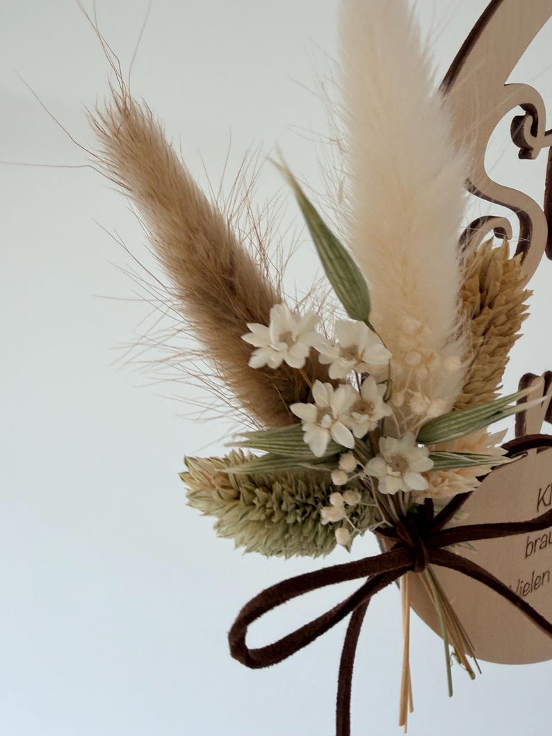 May include: A wooden wedding cake topper featuring a bouquet of dried flowers in beige, cream, and green hues. The bouquet is tied with a brown ribbon. The wooden base has decorative cutouts and text.