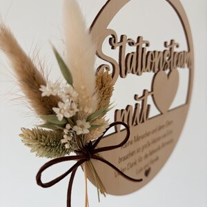 May include: A wooden circular sign with the words "Stationstraum mit" and a heart cutout. The sign is adorned with dried flowers and tied with a brown ribbon. A brown leather cord is attached for hanging.
