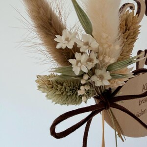 May include: A close-up of a dried flower arrangement with a brown suede ribbon. The arrangement features beige, brown, and green dried flowers and small white flowers. A wooden tag with text is visible.