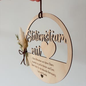 May include: A round wooden sign with the words "Stationsteam mit" cut out. The sign has a heart shape and a small bouquet of dried flowers and a leather cord for hanging. The sign also has text in German.