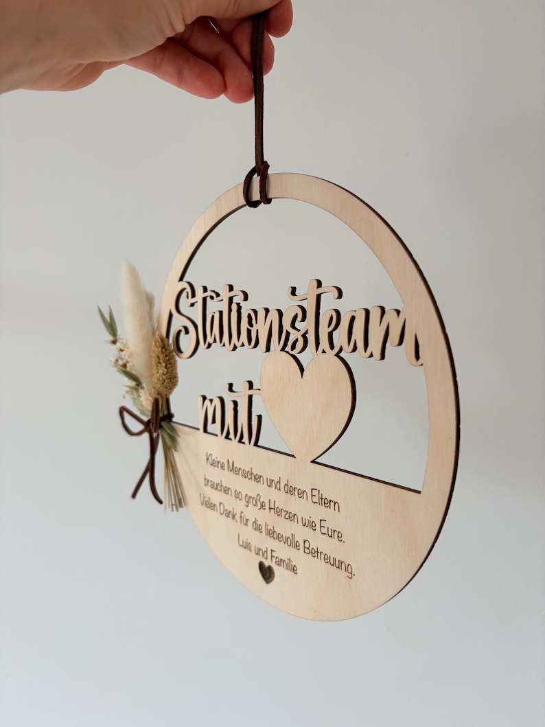 May include: A round wooden sign with the words "Stationsteam mit" cut out. The sign has a heart shape and a small bouquet of dried flowers and a leather cord for hanging. The sign also has text in German.