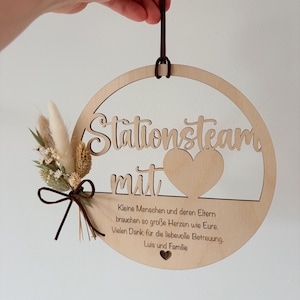 May include: A round wooden sign with the words "Stationsteam mit" cut out. The sign has a heart shape and a small bouquet of dried flowers and a brown ribbon. The sign has text in German.