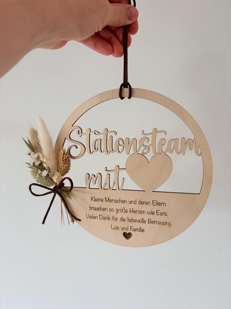 May include: A round wooden sign with the words "Stationsteam mit" cut out. The sign has a heart shape and a small bouquet of dried flowers and a brown ribbon. The sign has text in German.
