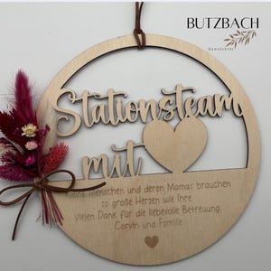 May include: Wooden round sign with the words "Stationsteam mit" cut out. A heart shape is in the center. A small bouquet of dried flowers in shades of pink and burgundy is tied to the side. The sign has a brown leather cord for hanging.
