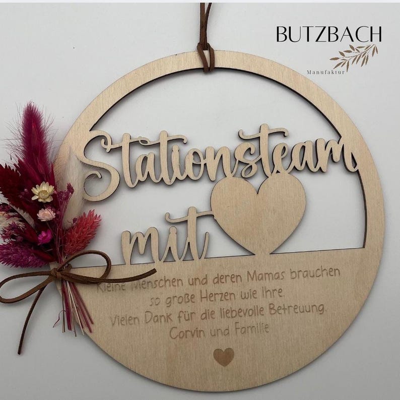 May include: Wooden round sign with the words "Stationsteam mit" cut out. A heart shape is in the center. A small bouquet of dried flowers in shades of pink and burgundy is tied to the side. The sign has a brown leather cord for hanging.