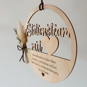 May include: A wooden circular sign with the words "Stationsteam mit" cut out. The sign has a heart shape and a small bouquet of dried flowers and a brown string for hanging. The sign also has text that reads "Kleine Menschen und deren Eltern brauchen so gro&szlig;e Herzen wie Eure. Vielen Dank f&uuml;r die liebevolle Betreuung. Luis und Familie."