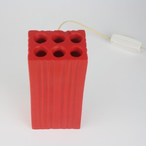 Pu&ograve; includere: Lampada da scrivania rossa, rettangolare, con sei aperture circolari sulla parte superiore. La lampada presenta scanalature verticali sui lati e un cavo color crema con una spina bianca. Lampada decorativa unica.