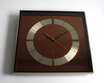 May include: A square wall clock featuring a metallic gold frame and a deep brown face. A circular gold dial with black Roman numeral markers sits in the center. The clock has ornate black hour and minute hands and a thin gold second hand.