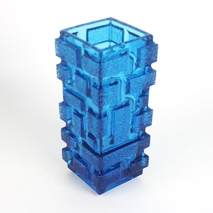 Pode incluir: Um vaso de vidro azul de formato quadrado com um padrão geométrico texturizado semelhante a tijolos no exterior. O recipiente tem uma aparência translúcida com vários tons de azul criados pela profundidade do vidro moldado.