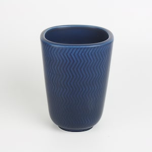Può includere: Un bicchiere in ceramica blu navy con un motivo a zig-zag strutturato. La tazza cilindrica ha una base arrotondata e un bordo liscio di forma ovale. Il colore blu scuro e il design a zig-zag conferiscono al bicchiere un aspetto moderno.