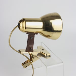Puede incluir: Una lámpara dorada con clip, con soporte marrón y cable curvado. La lámpara tiene una pantalla redondeada y una base de clip para fijar. El diseño es vintage y funcional.