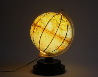 Puede incluir: Una lámpara de globo vintage iluminada. El globo muestra un mapa detallado del mundo, iluminado desde el interior. La lámpara tiene una base negra y un marco de metal. El globo tiene aproximadamente 30 cm de diámetro.