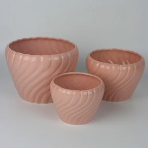 May include: Three pink ceramic planters of varying sizes. Each planter has a wavy, textured design and a scalloped rim. The planters are a soft, muted pink color, suitable for indoor or outdoor use.