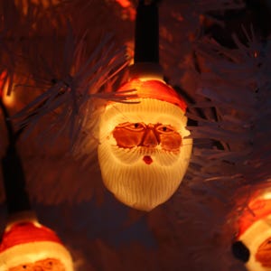 May include: Close-up of Christmas lights shaped like Santa Claus faces. Each light has a red hat, a white beard, and a warm, glowing light. The lights are strung on a dark wire, and the background is a white Christmas tree.