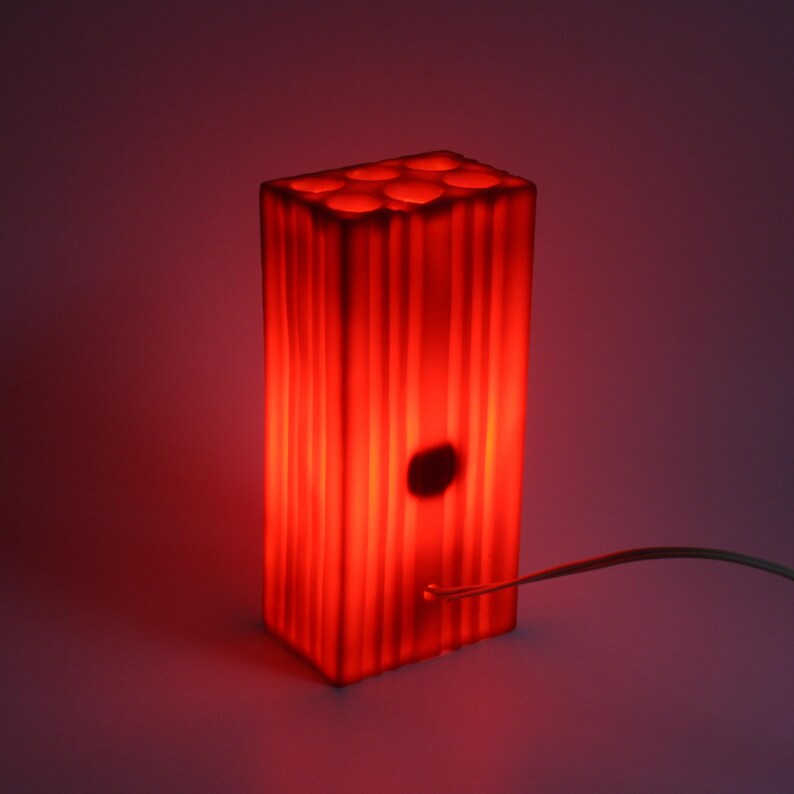Pu&ograve; includere: Lampada rettangolare rossa con linee verticali e aperture circolari sulla parte superiore. La lampada emette una calda luce rossa, con un elemento circolare scuro sul davanti. Un sottile cavo si estende dalla base.