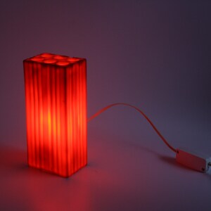 Pu&ograve; includere: Una lampada rettangolare rossa con linee verticali, che emette un bagliore caldo. La lampada ha aperture circolari sulla parte superiore. Un adattatore di alimentazione bianco &egrave; collegato alla lampada tramite un sottile filo arancione.