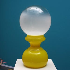 May include: A decorative glass object with a spherical top and a yellow base. The top is a clear sphere that fades to white. The base is a bright yellow color with a bulbous shape. The object is set against a blue background.