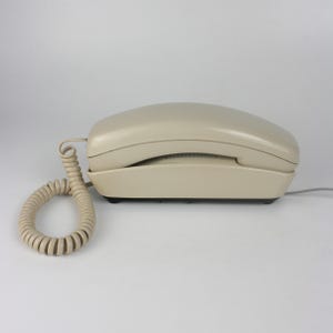 May include: A beige landline telephone with a smooth, curved handset resting on a matching base. The device features a coiled cord extending from the side of the handset to the base unit, which has a dark brown bottom and a thin grey cord extending out.
