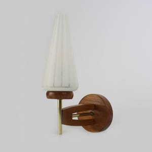 Può includere: Una lampada da parete con paralume conico in vetro bianco a coste verticali montato su una base circolare in legno. L'apparecchio include uno stelo in metallo color ottone che collega il paralume a un braccio in legno fissato alla parete.