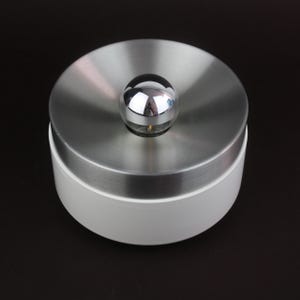 May include: A contemporary, circular container featuring a white base and a brushed metal lid. A small, chrome sphere is positioned on top of the lid. The container is likely for storage or decorative use.