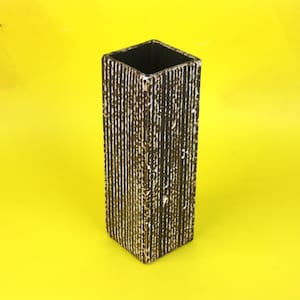 May include: A tall, rectangular vase with a textured surface. The vase is primarily dark brown with vertical white and gold accents. The interior of the vase is black, and it sits against a bright yellow background.