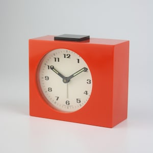 May include: A vibrant orange, square alarm clock with a white clock face and black numerals. The clock features black hands and a small black square on top. The clock face is approximately 7.6 cm in diameter.
