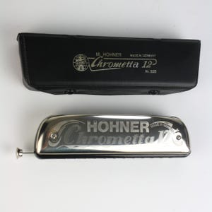 May include: A silver Hohner Chrometta 12 harmonica with a black case. The harmonica has the words "Hohner Chrometta 12" engraved on it. The black case has the words "M. Hohner Chrometta 12" and "Made in Germany" printed on it.